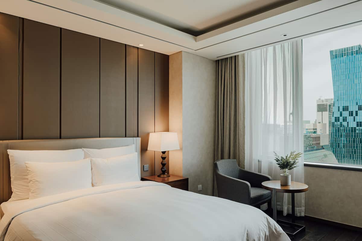 A hotel room with a large bed, a bedside table with a lamp, a chair, and a small table with a plant. The room has a view of a cityscape through large windows.