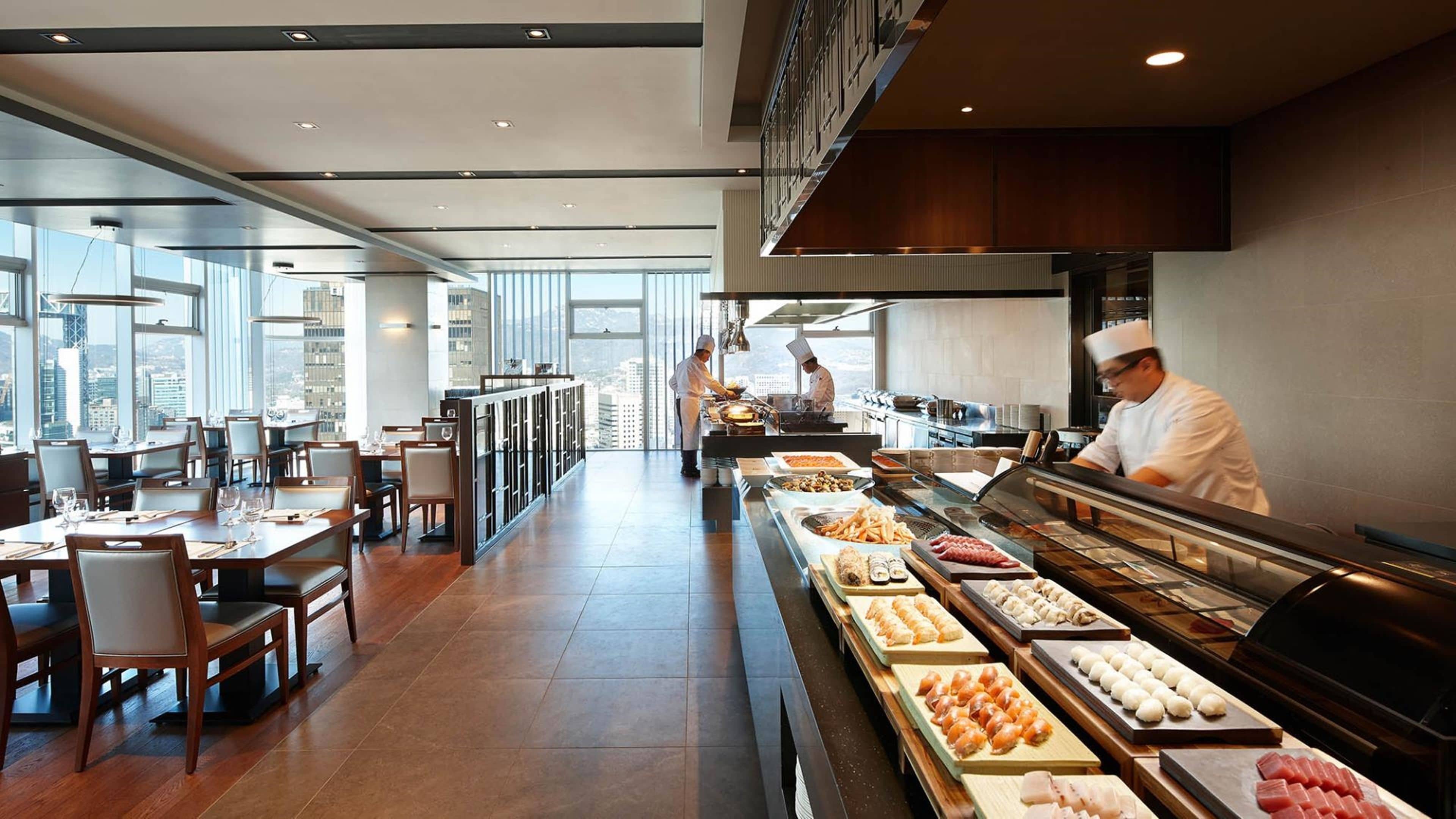 A modern restaurant with a long counter displaying various dishes, a dining area with tables and chairs, and large windows offering a city view.