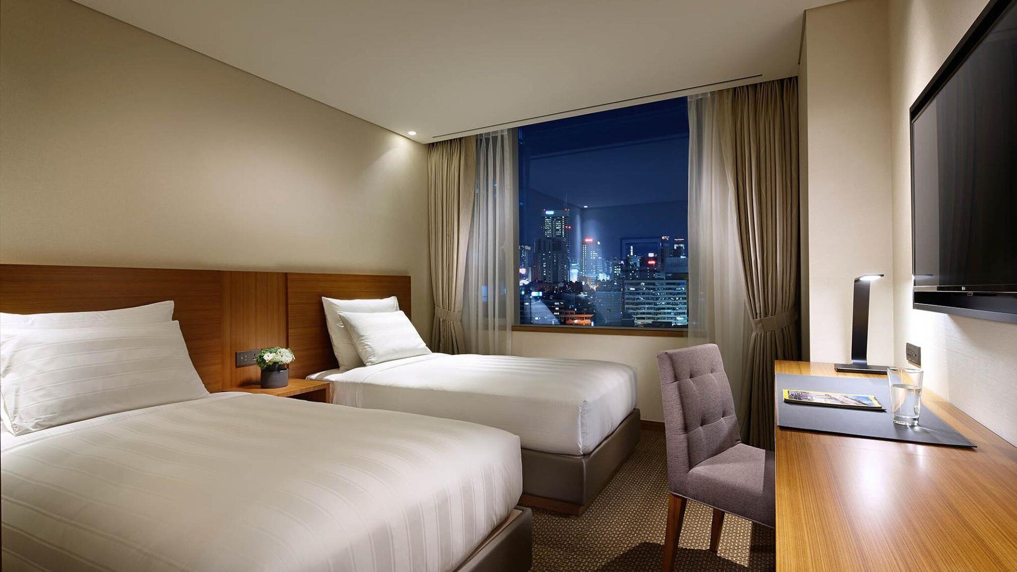 Habitación de hotel con dos camas, un escritorio y una silla, televisión y vista a un paisaje urbano nocturno.