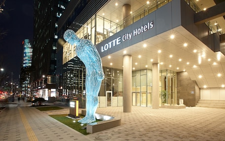 A modern hotel entrance with a prominent geometric sculpture, a well-lit facade, and a cityscape in the background.