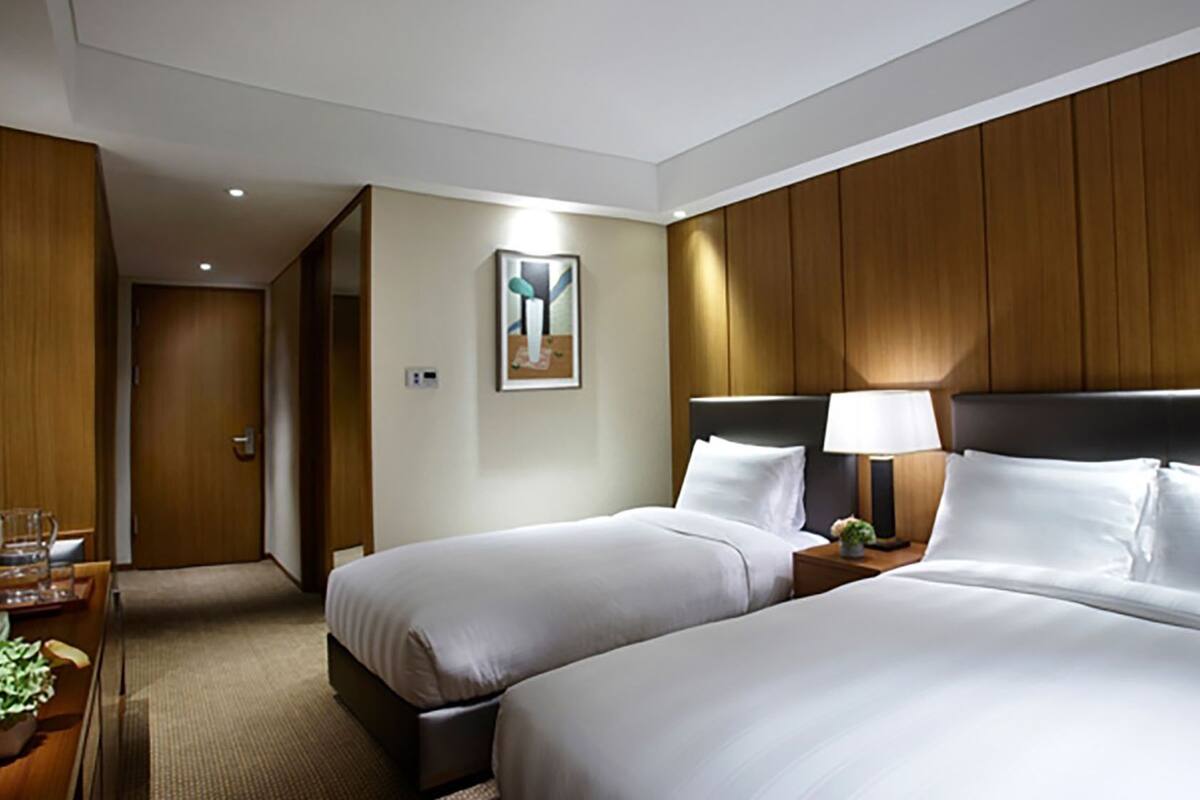 A hotel room with two beds, a nightstand with a lamp, a small table with a glass of water, and a painting on the wall.