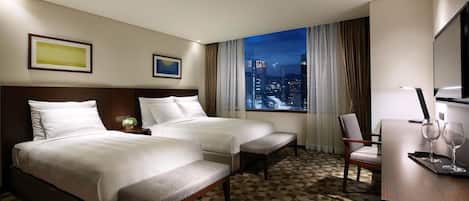Habitación de hotel con dos camas, un escritorio con silla, televisión y vista a la ciudad.