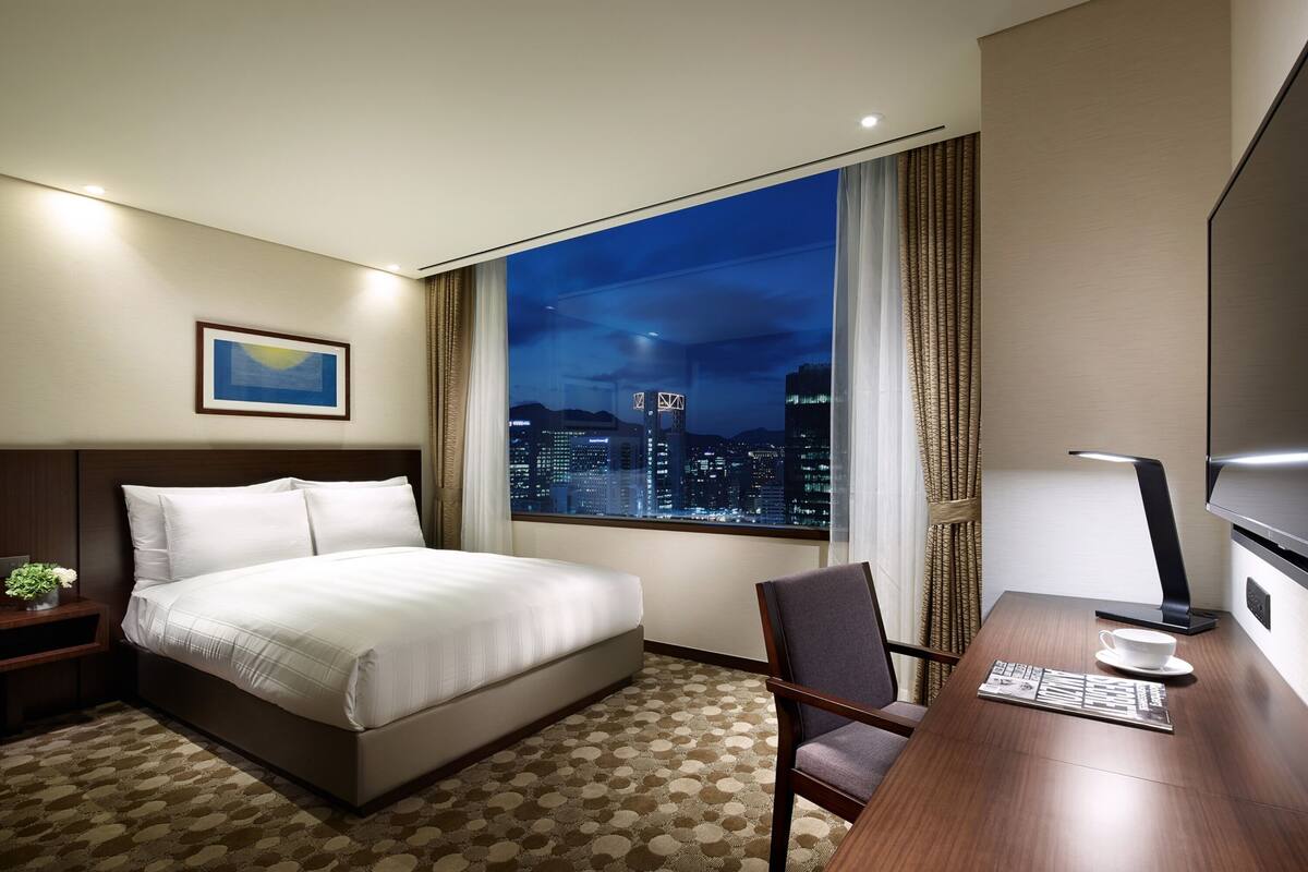 A hotel room with a large bed, a desk with a chair, a window with a city view, and a framed picture on the wall.