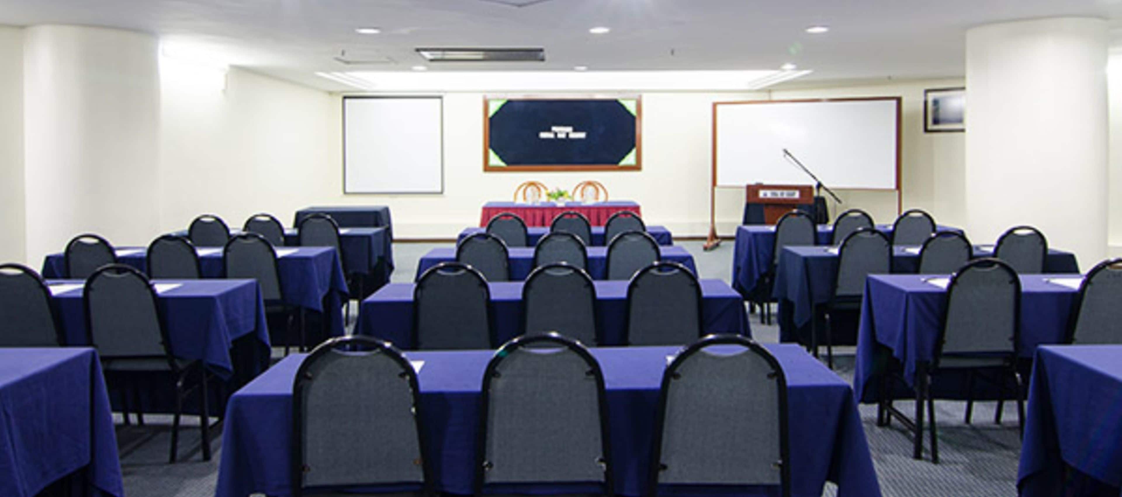 A conference room with rows of tables and chairs, a podium, and two projection screens.