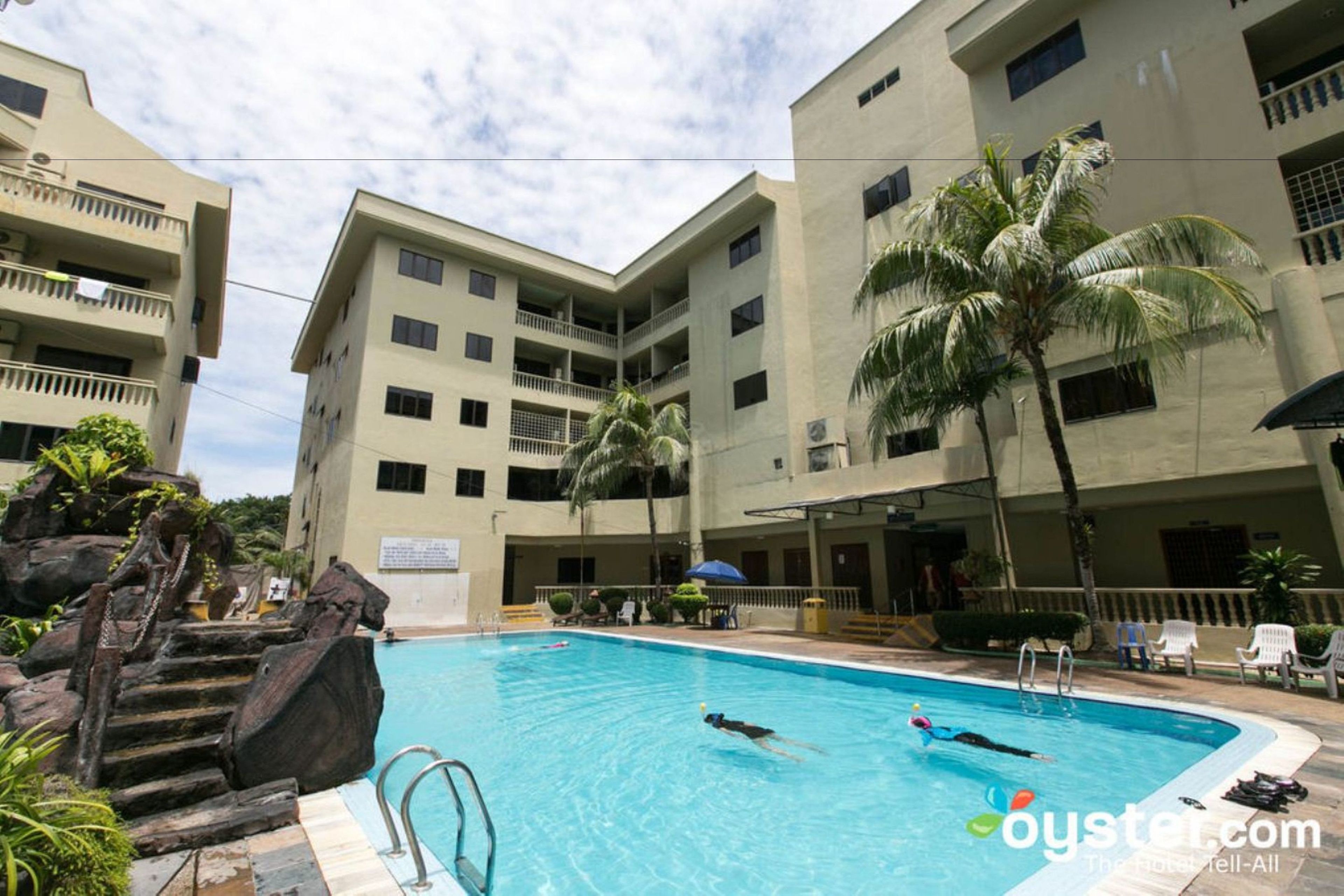A swimming pool surrounded by multi-story buildings with balconies, palm trees, and a stone staircase.