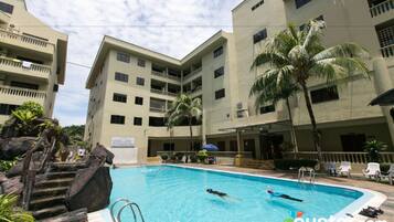 A swimming pool surrounded by multi-story buildings with balconies, palm trees, and a stone staircase.