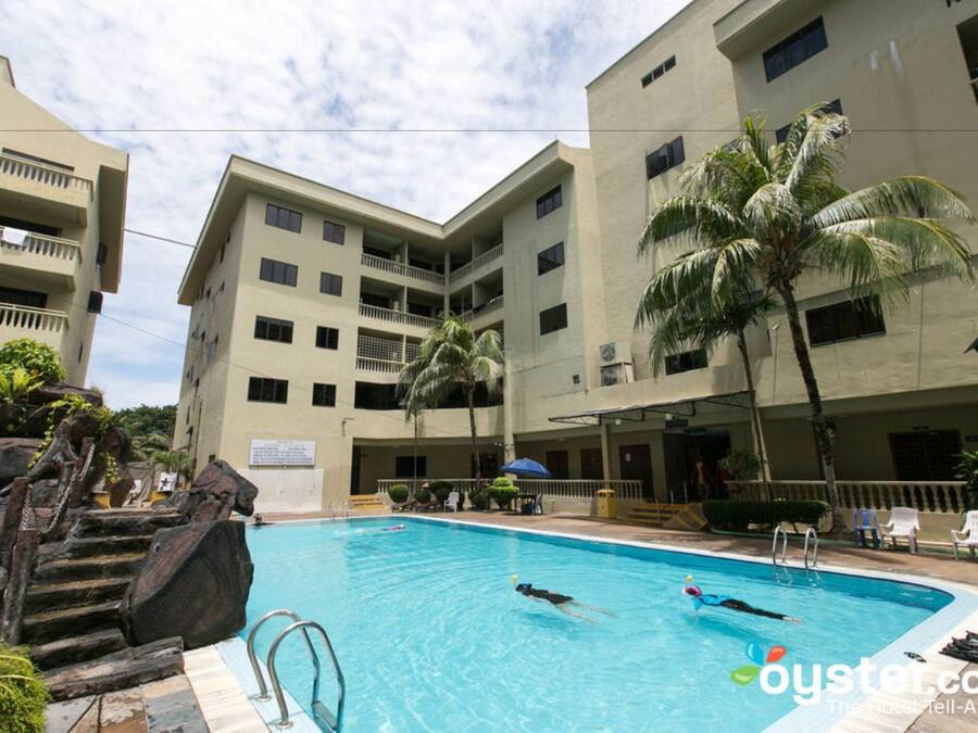 A swimming pool surrounded by multi-story buildings with balconies, palm trees, and a stone staircase.