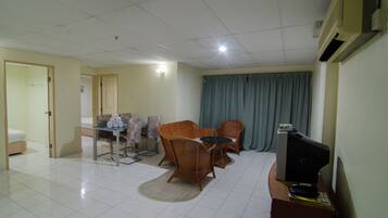 Una habitación con una mesa de comedor, dos sillas de mimbre, un televisor pequeño sobre un mueble bajo y una cama al fondo.