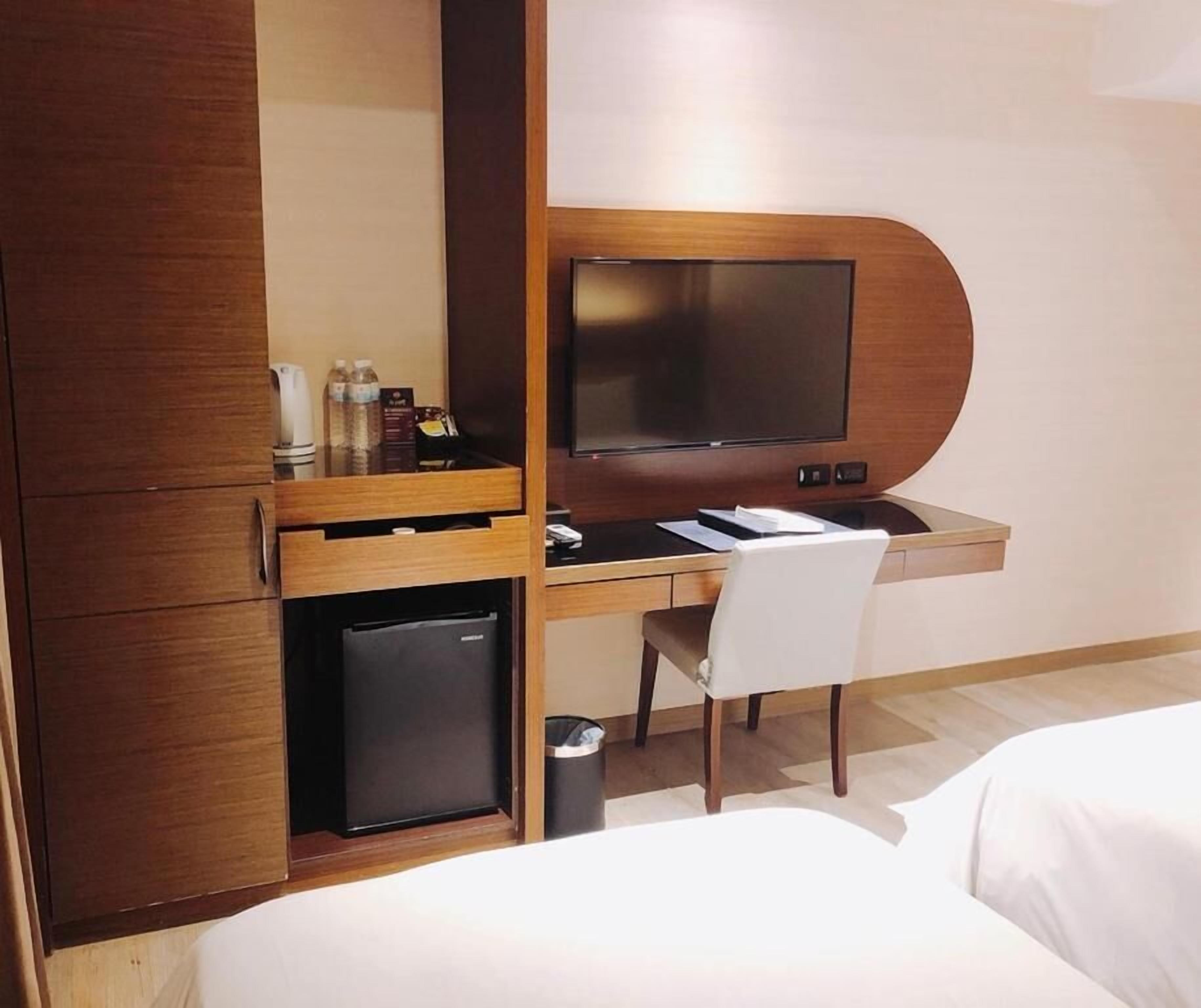 A hotel room with a wooden desk, a television, a small refrigerator, a chair, and a bed.