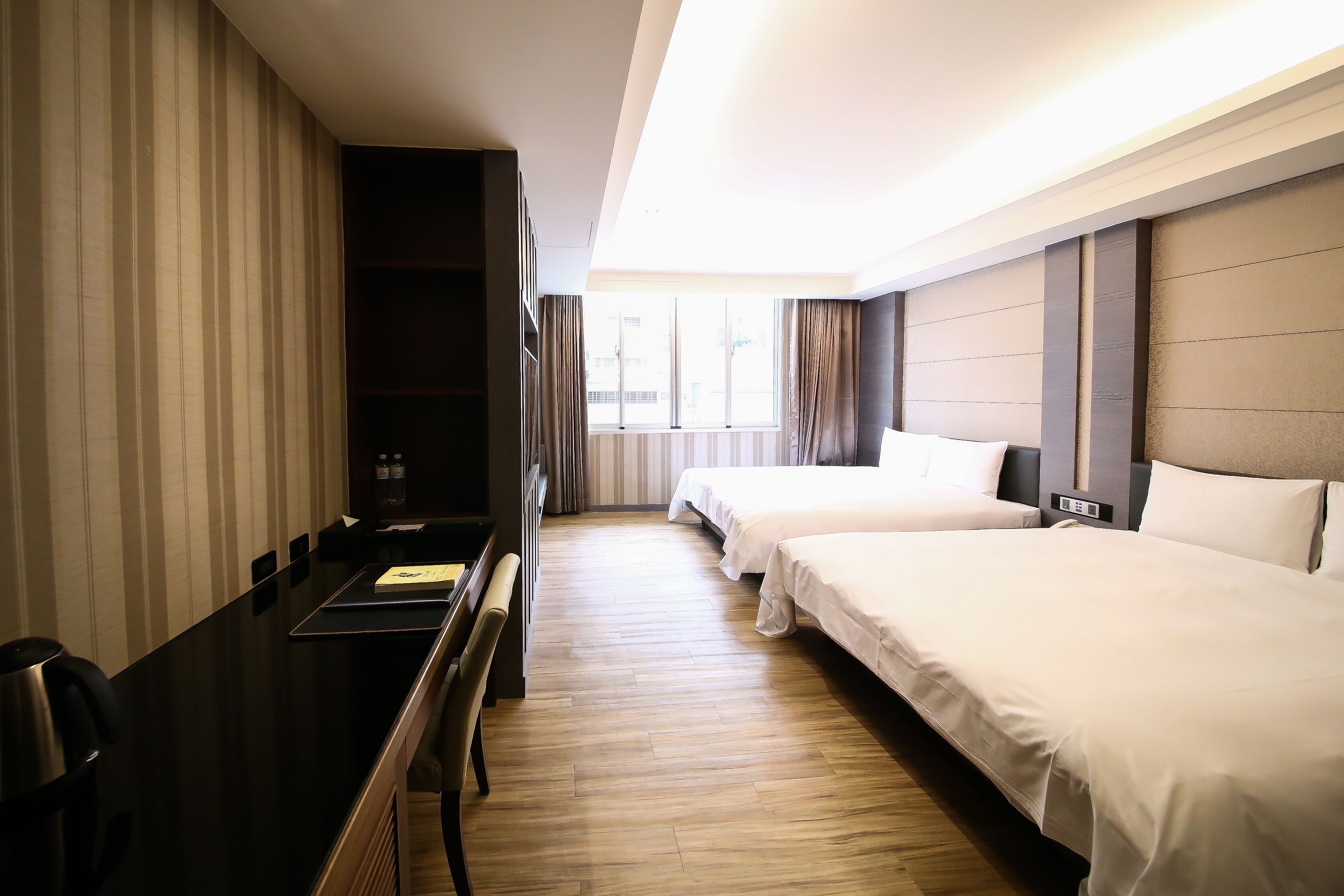 A hotel room with a desk, two beds, a window, and a ceiling light.