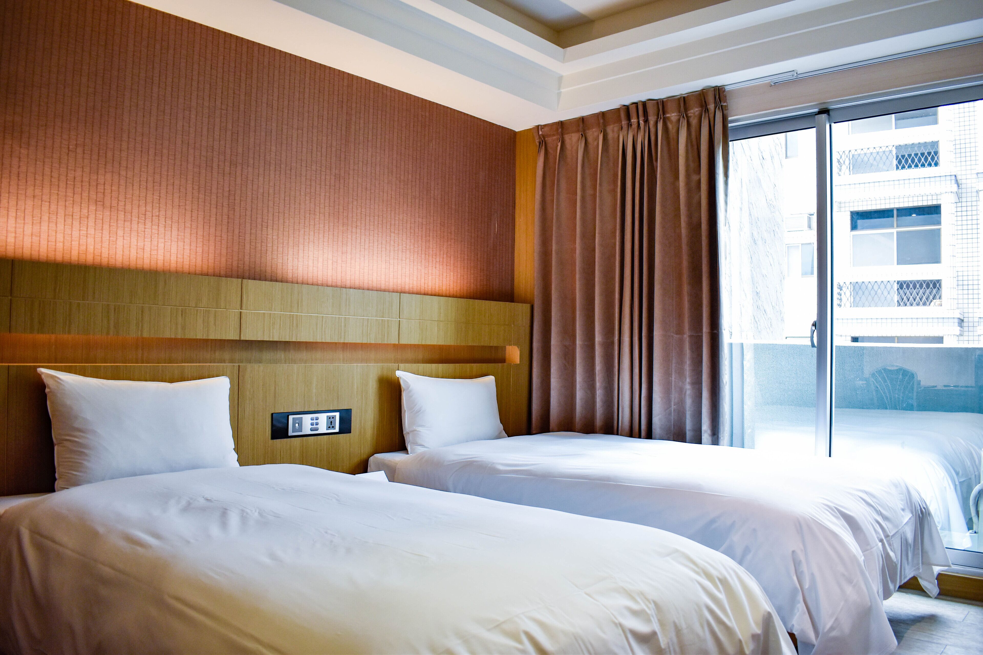 A hotel room with two beds, a wooden headboard, a large window with curtains, and a view of the city.