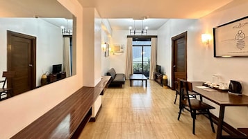 Una habitación de hotel moderna con suelo de madera, un sofá, una mesa de comedor con sillas, un espejo grande y vistas a la ciudad desde la ventana.