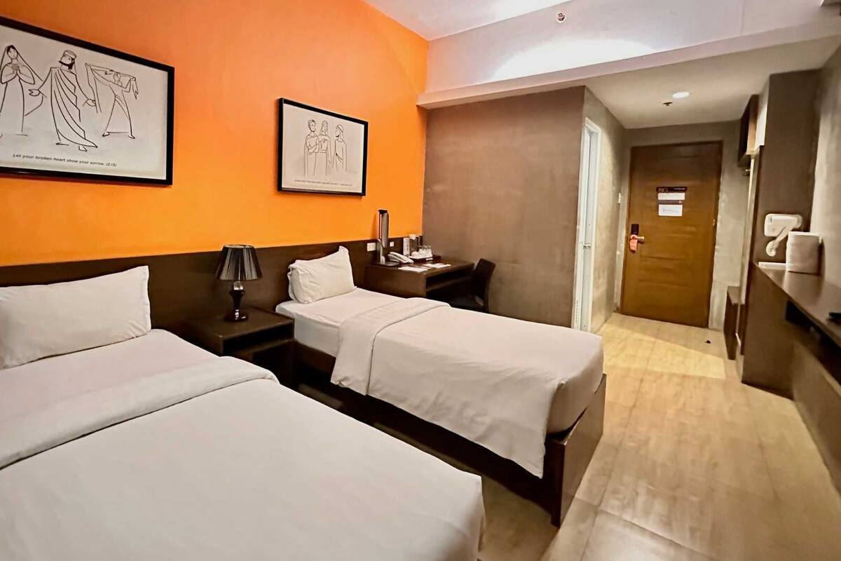 Una habitación de hotel con dos camas, una mesita de noche con lámpara, un cuadro enmarcado en la pared y una puerta que da a otra habitación.