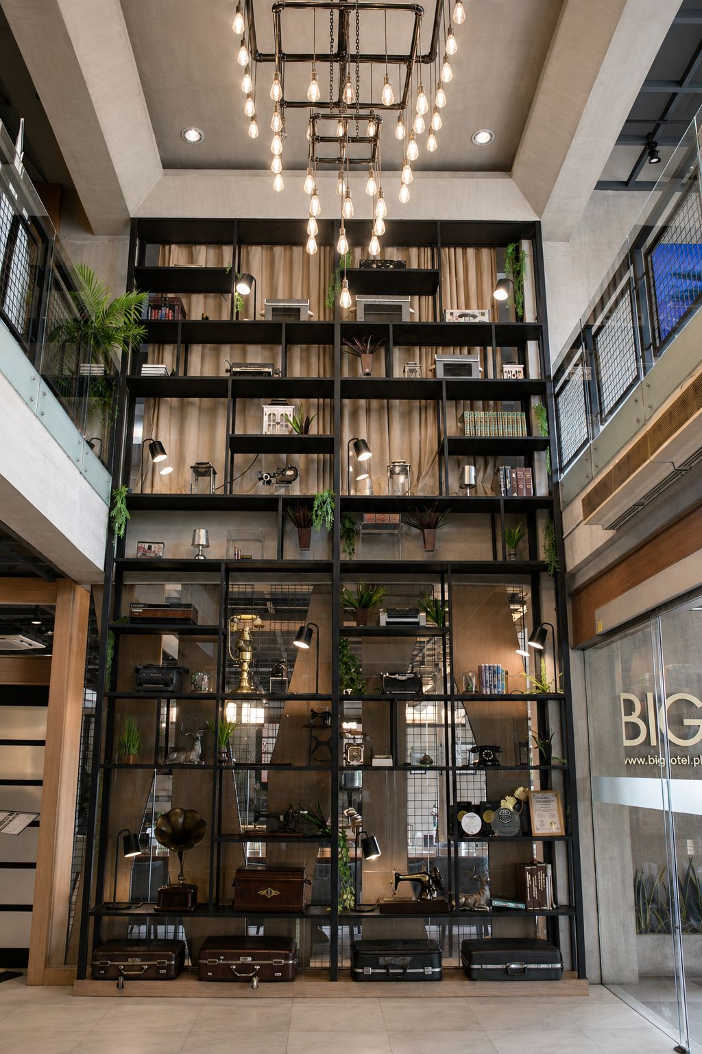 A lobby with a modern design, featuring a multi-level black shelving unit with various items, a central hanging light fixture, and a visible entrance to a hotel named 'BIG'.