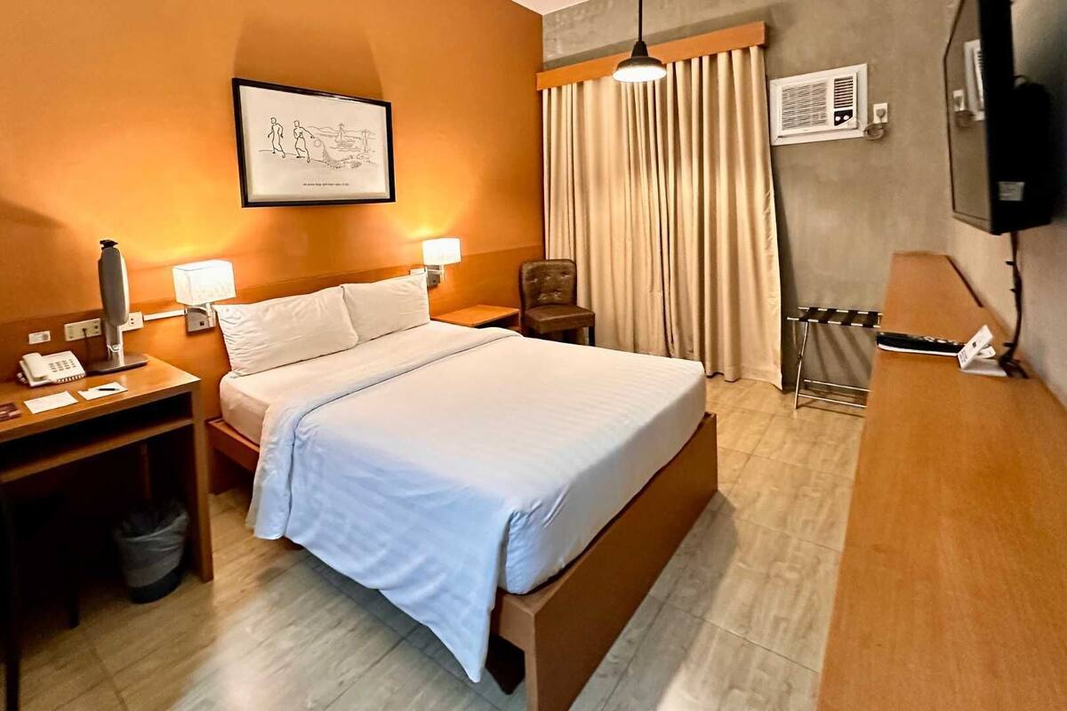 Habitación de hotel con cama, mesitas de noche, escritorio con teléfono, silla, cuadro en la pared y televisor.