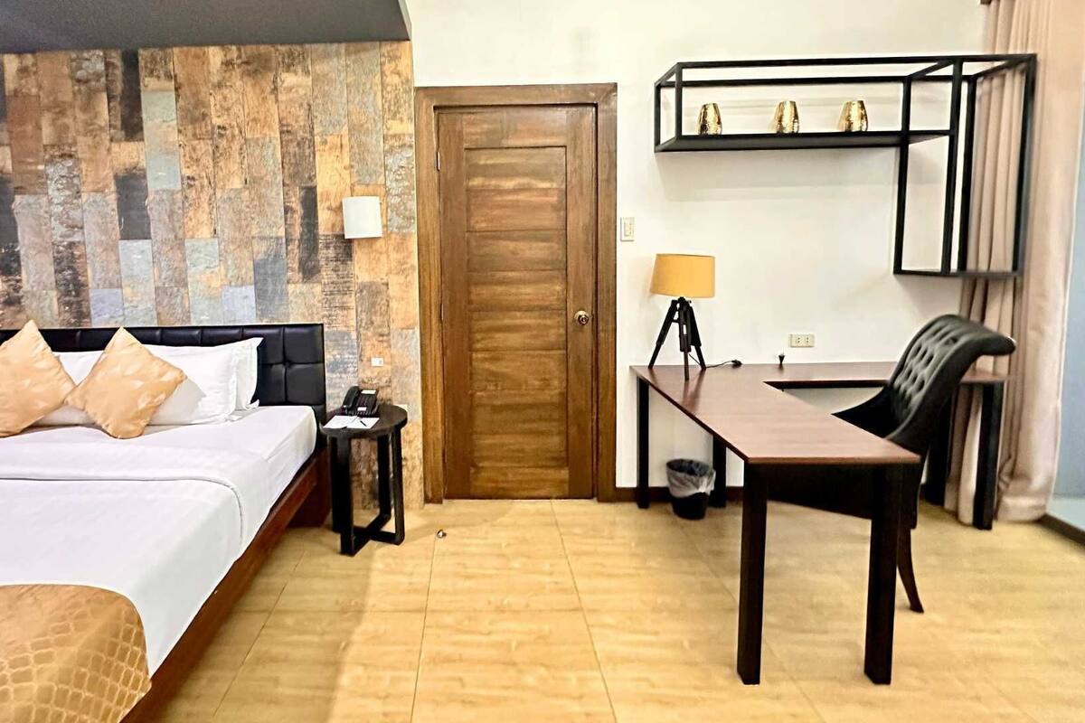 Una habitación de hotel con cama, mesita de noche, lámpara, escritorio, silla y una puerta de madera.