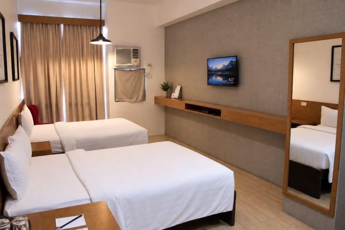 A hotel room with two beds, a desk with a chair, a mounted TV, a mirror, and a window with curtains.