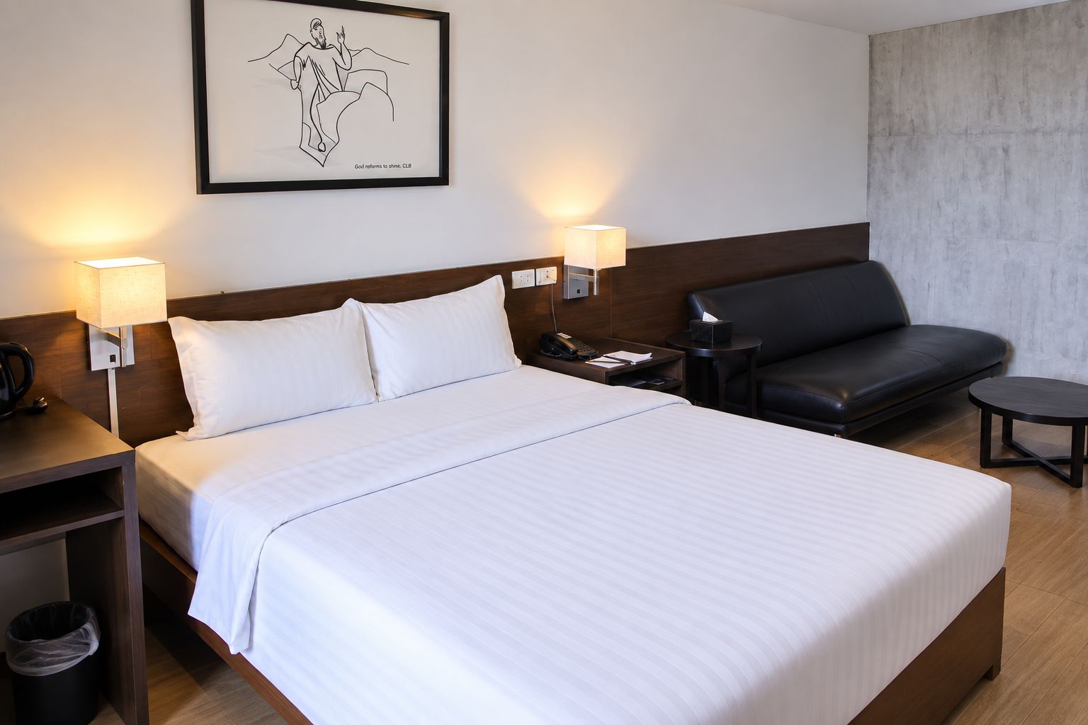 A hotel room with a large bed, two bedside lamps, a black leather sofa, a small round table, and a framed artwork on the wall.