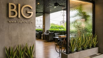 A modern hotel entrance with a large sign reading 'BIG HOTEL' and a website address. There are indoor seating areas with tables and chairs, and a bicycle parked outside.