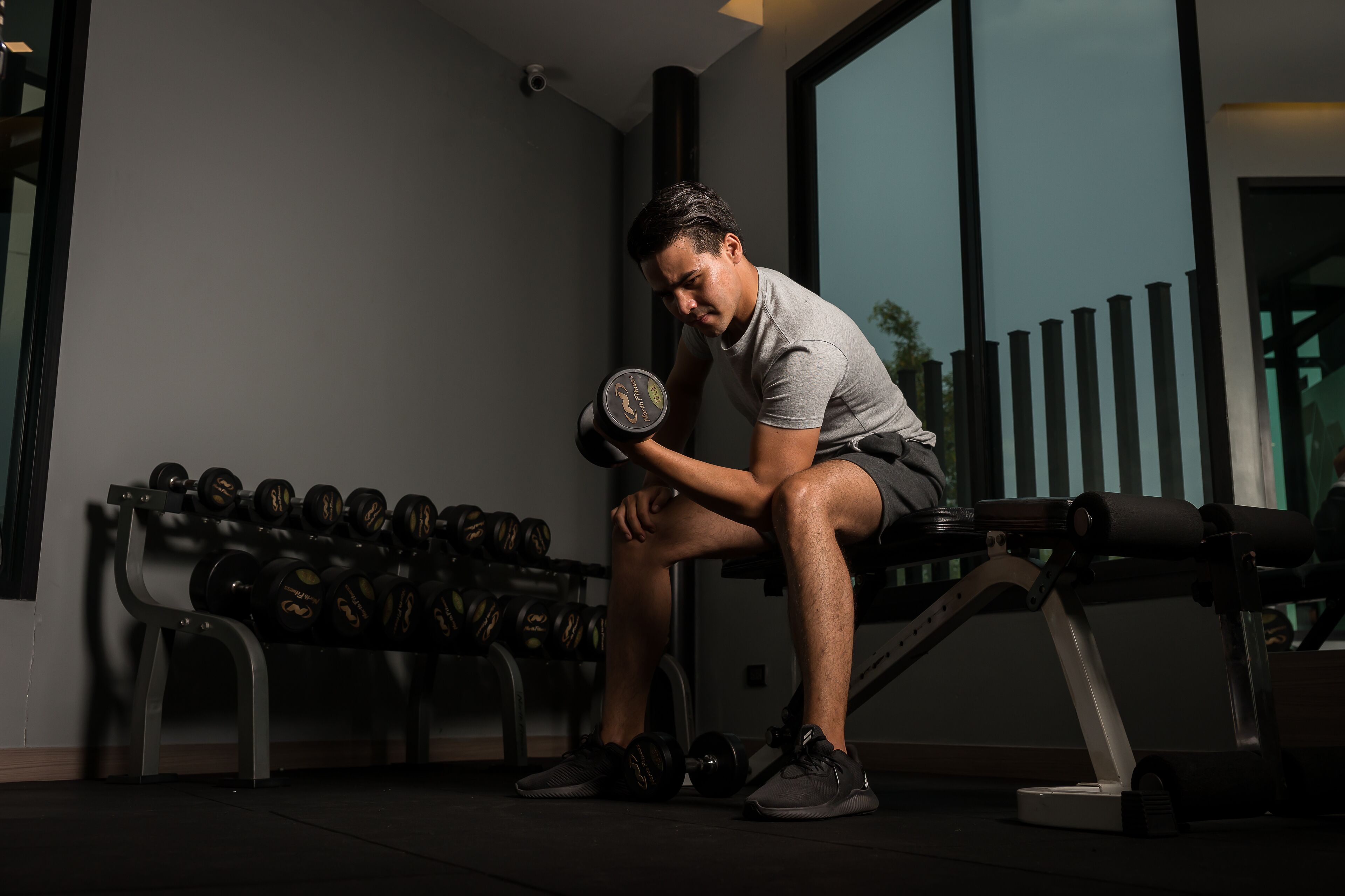 Un gimnasio con una persona sentada en un banco, sosteniendo una pesa. Se ven varias pesas en un soporte al fondo.