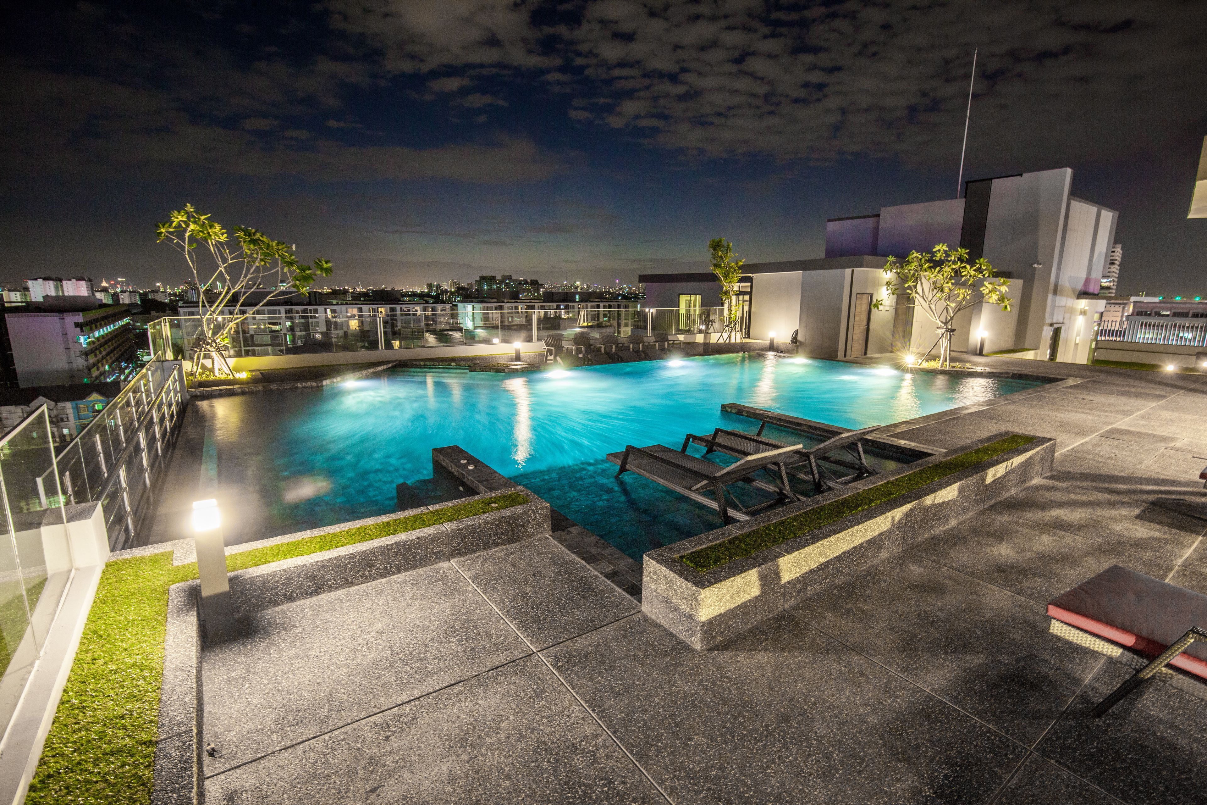 Une piscine sur le toit offrant une vue dégagée sur les gratte-ciel de la ville la nuit, entourée d’une architecture moderne et dotée de chaises longues et d’un jacuzzi.