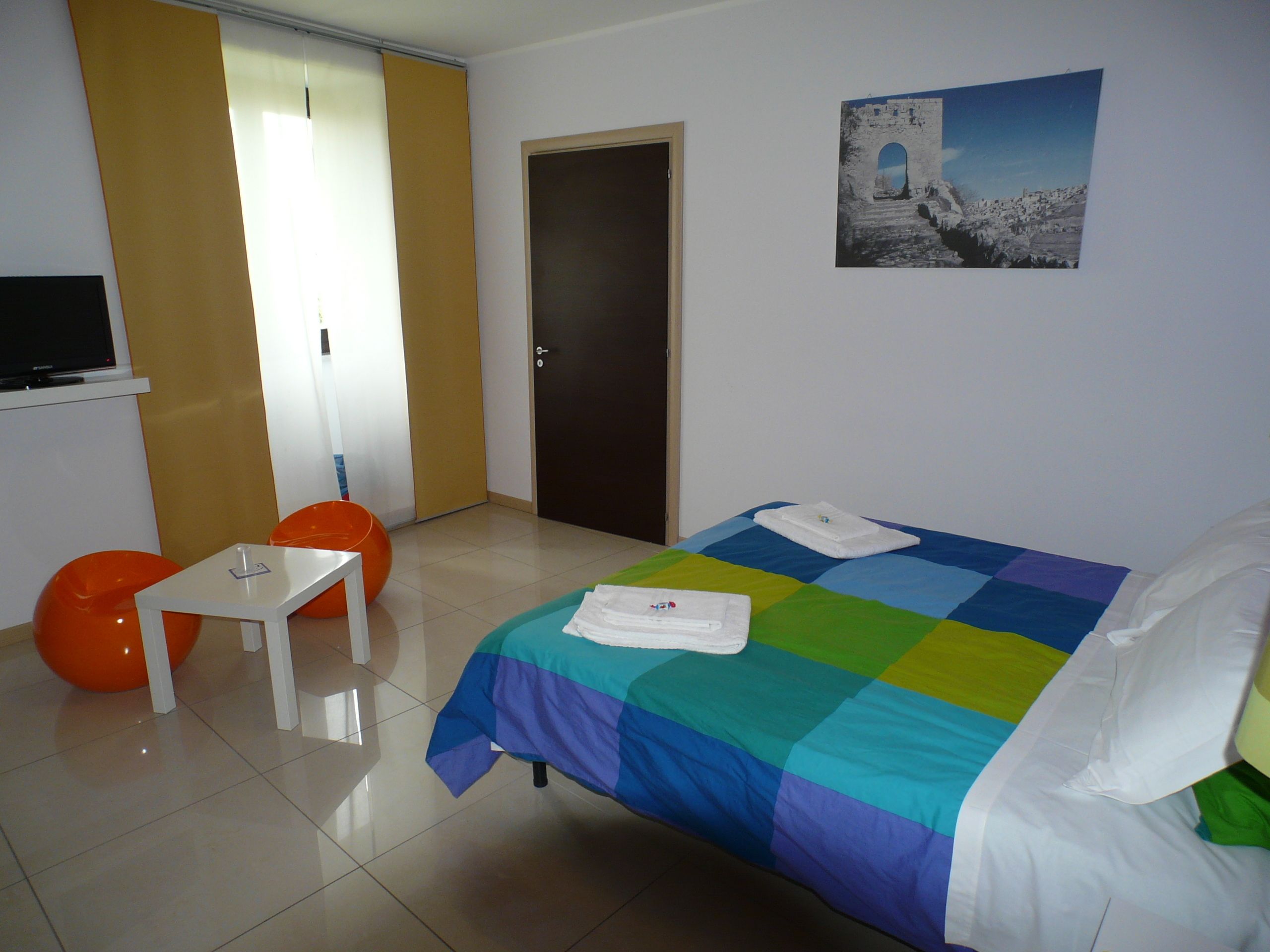 A hotel room with a bed, a small table, two orange chairs, a television, and a door.
