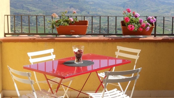 Uma varanda com uma mesa vermelha e cadeiras brancas, com vista para uma paisagem montanhosa.