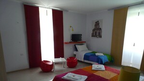 Quarto de hotel com uma cama, televisão, uma cadeira vermelha, uma mesinha e uma janela com cortinas.