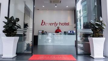 The reception area of the Beverly Hotel with a receptionist behind the desk, a red carpet, and potted plants on either side.