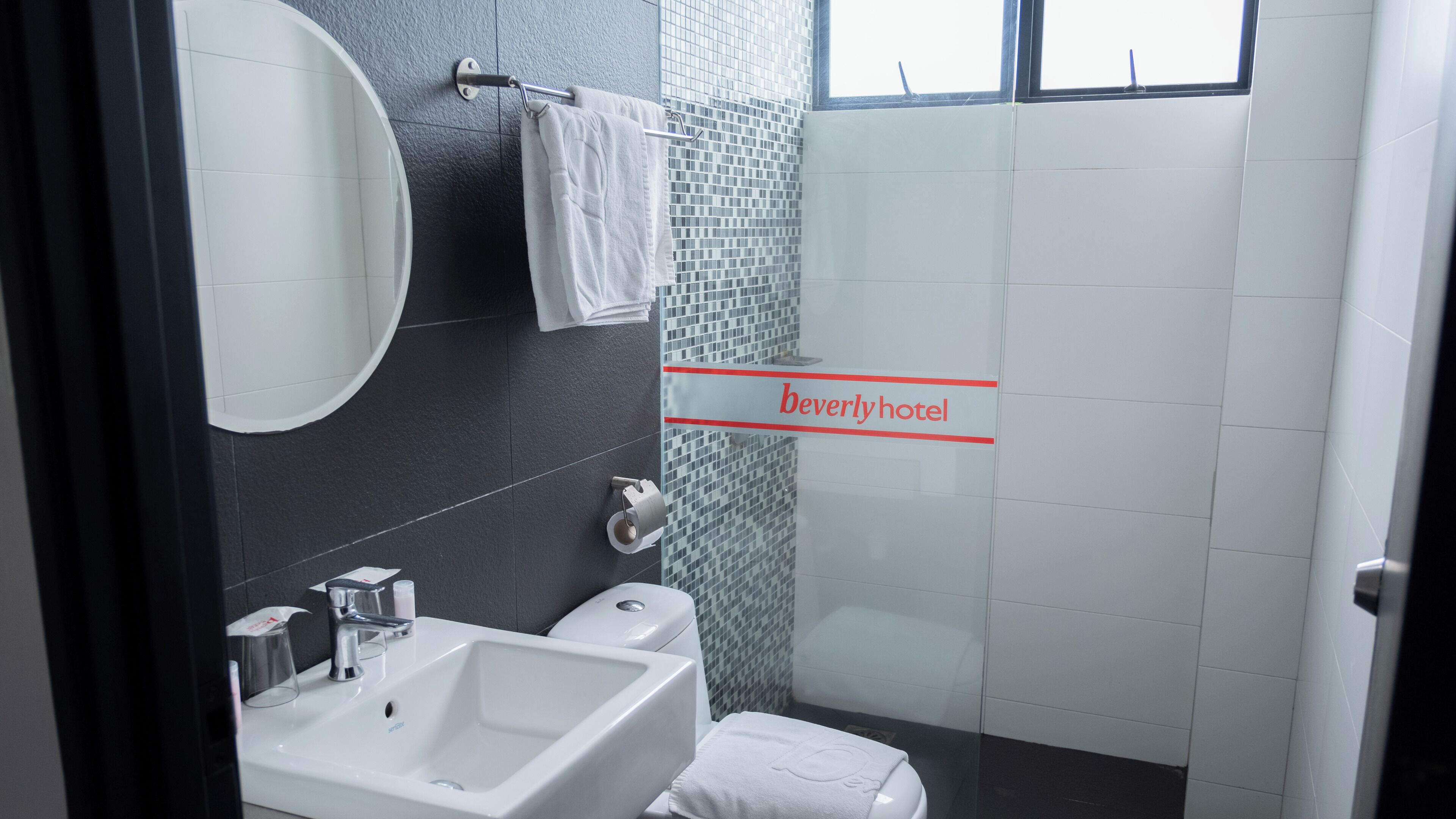 A bathroom with a sink, mirror, and toilet. There is a towel rack with a towel hanging on it. The walls are tiled, and there is a window above the shower area.