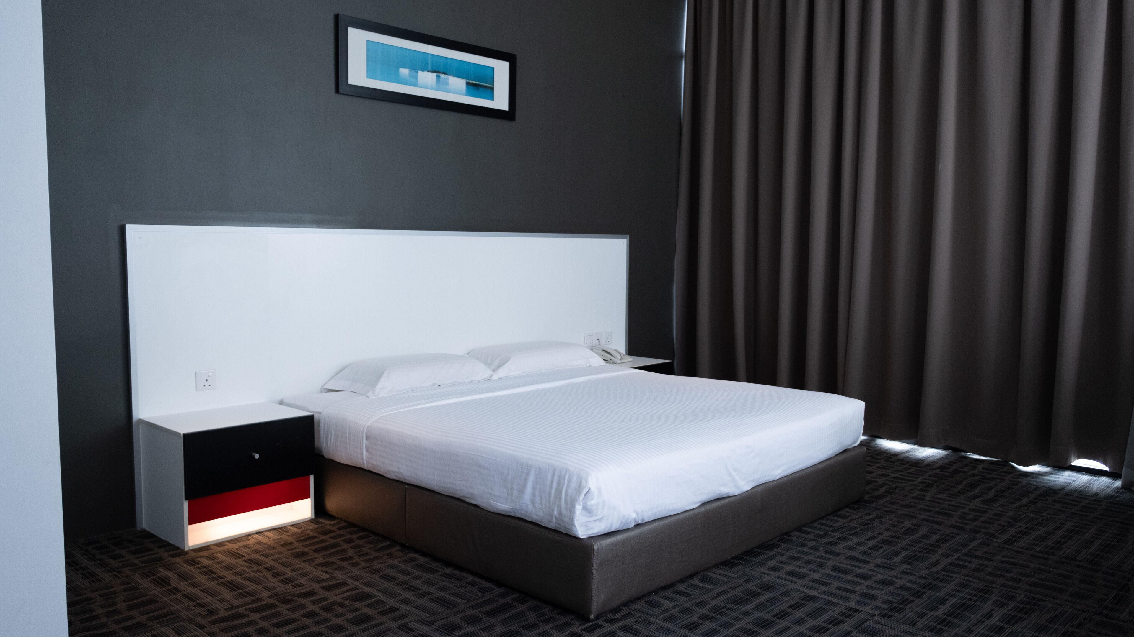 A hotel room with a large bed, a bedside table, a power outlet, and a framed picture on the wall.