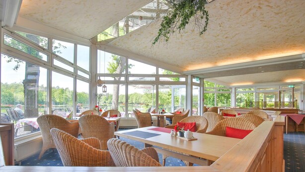 A restaurant with wicker furniture, large windows, and a view of greenery. - Emsland Hotel Saller See (Freren)