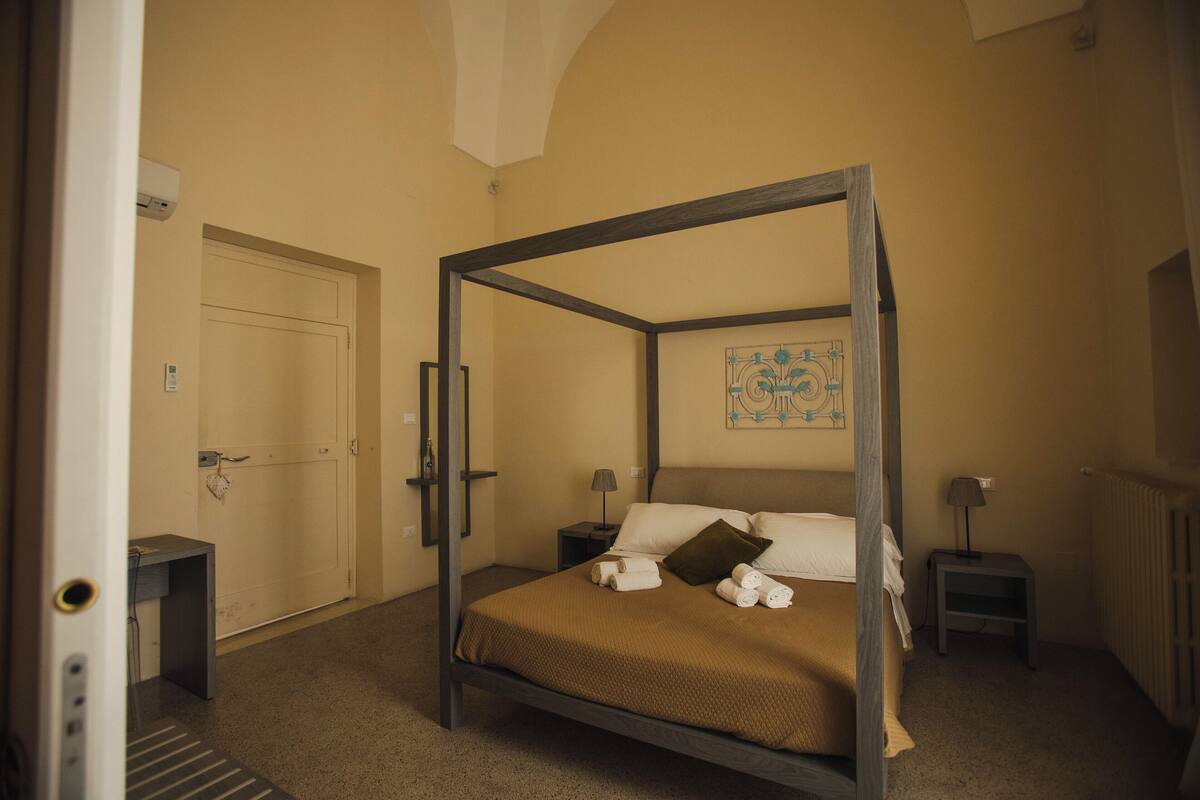 A four-poster bed with a brown bedspread, two bedside tables with lamps, a door, and a wall-mounted air conditioning unit.