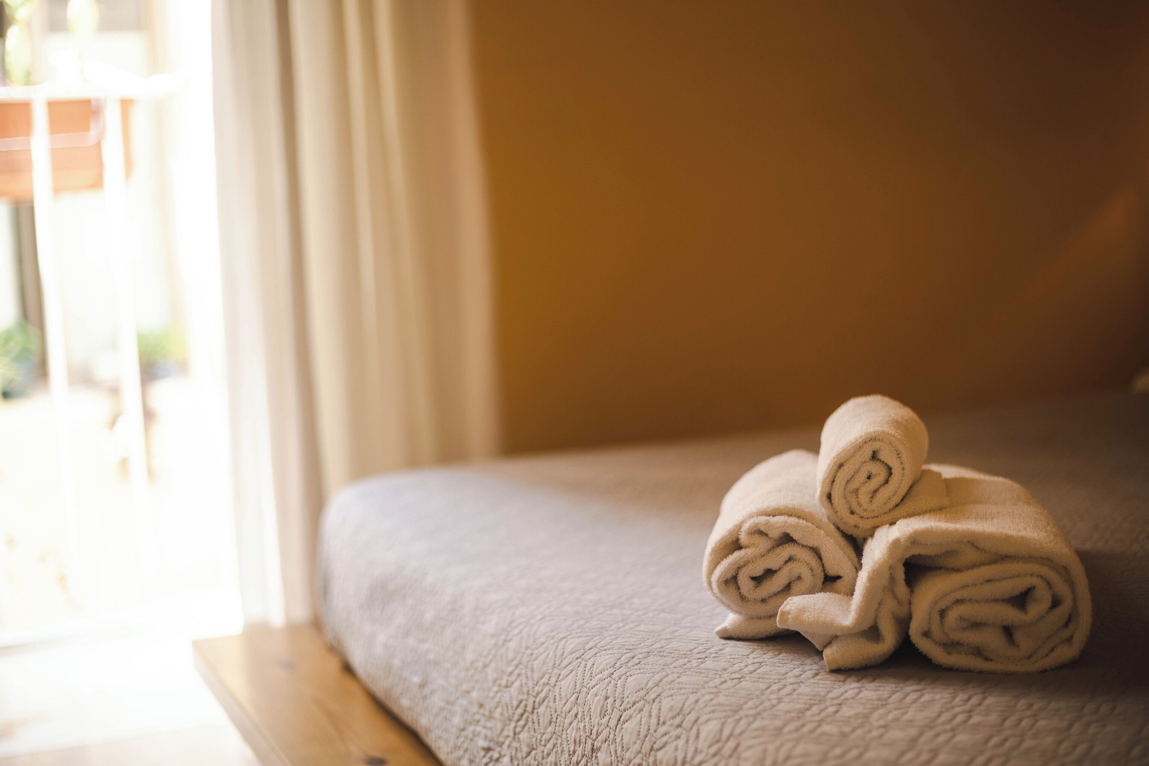 A neatly made bed with rolled towels on the side, a wooden bench, and a window with curtains.