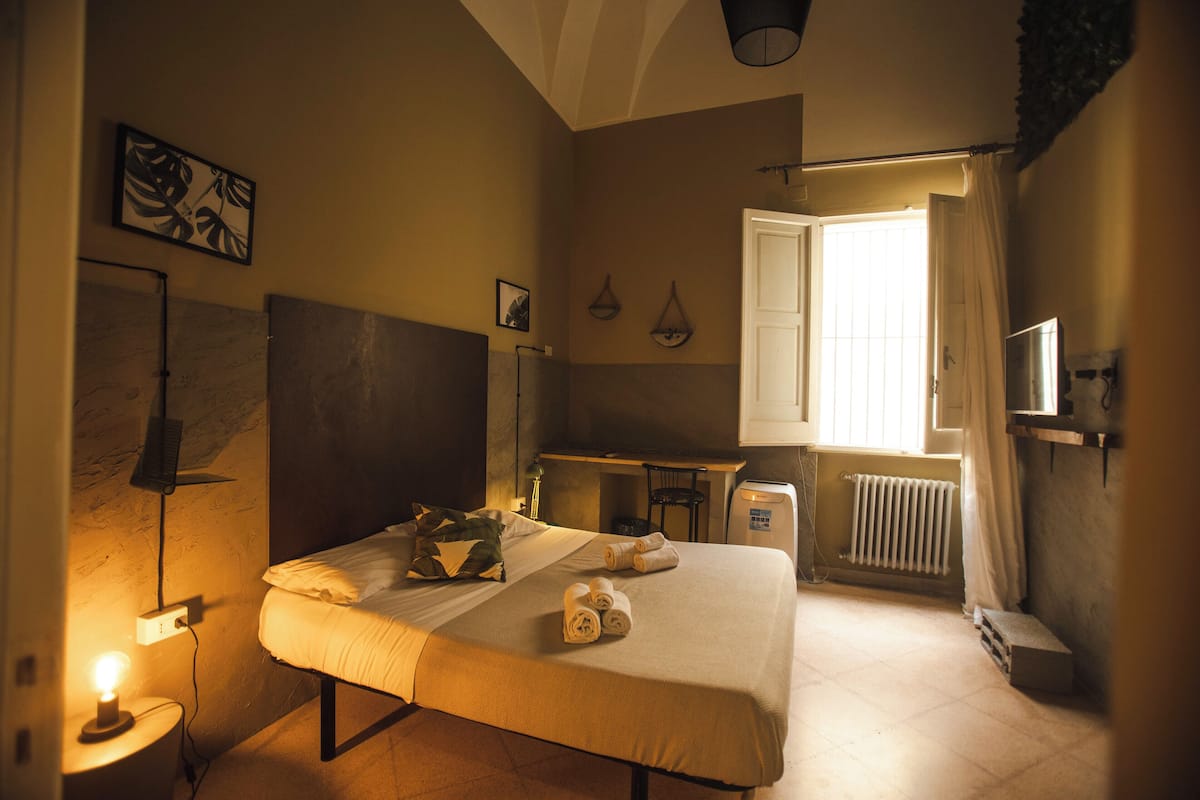 A hotel room with a bed, bedside lamp, a desk, a chair, a radiator, and a television.