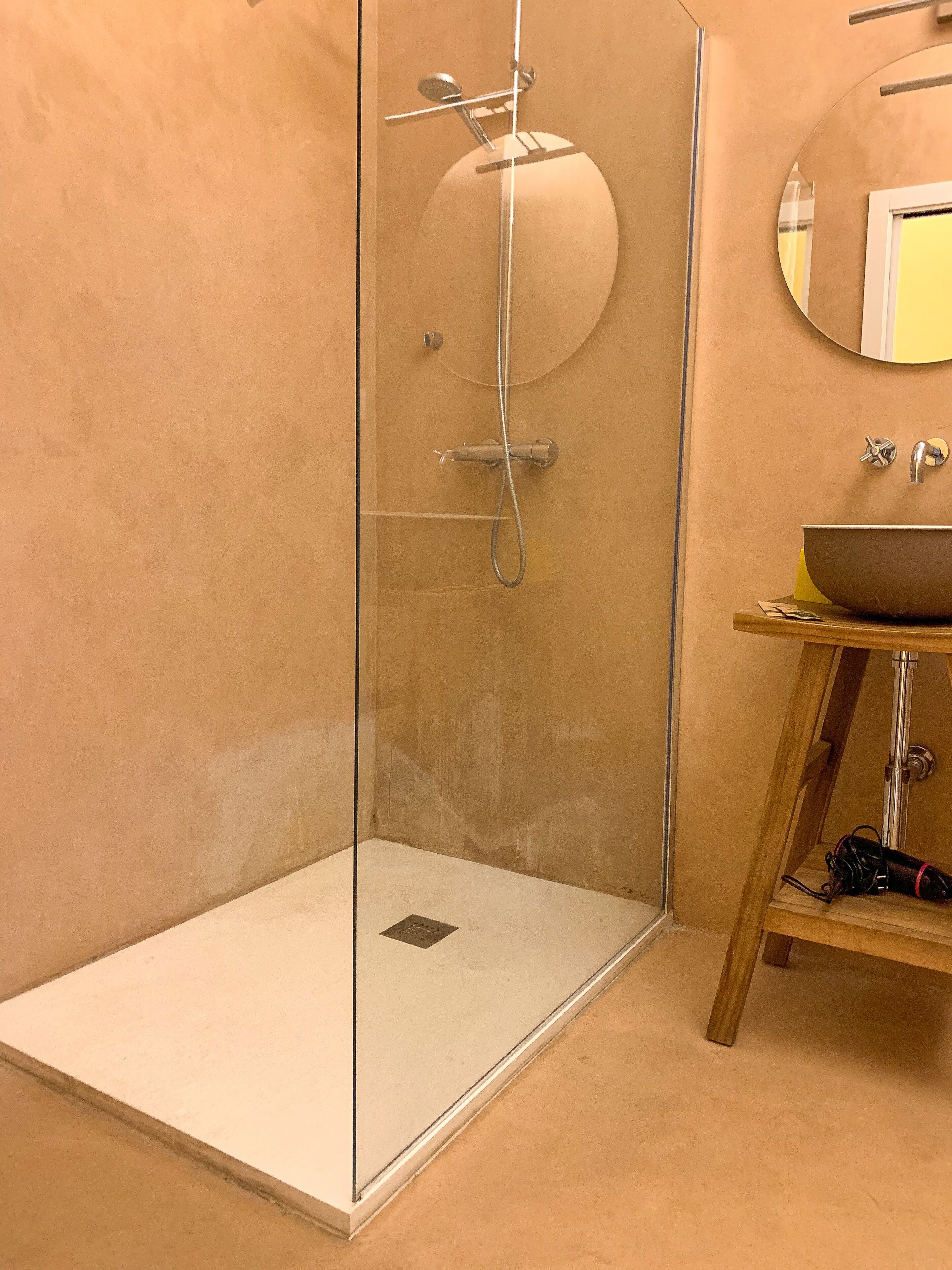 Ducha con cerramiento de vidrio, cabezal de ducha de mano, espejo redondo y taburete de madera con lavabo.