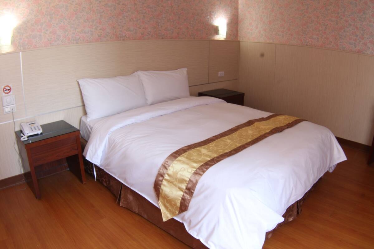 Cama bem arrumada com edredom branco e castanho, mesa de cabeceira com telefone e luminária fixada na parede.