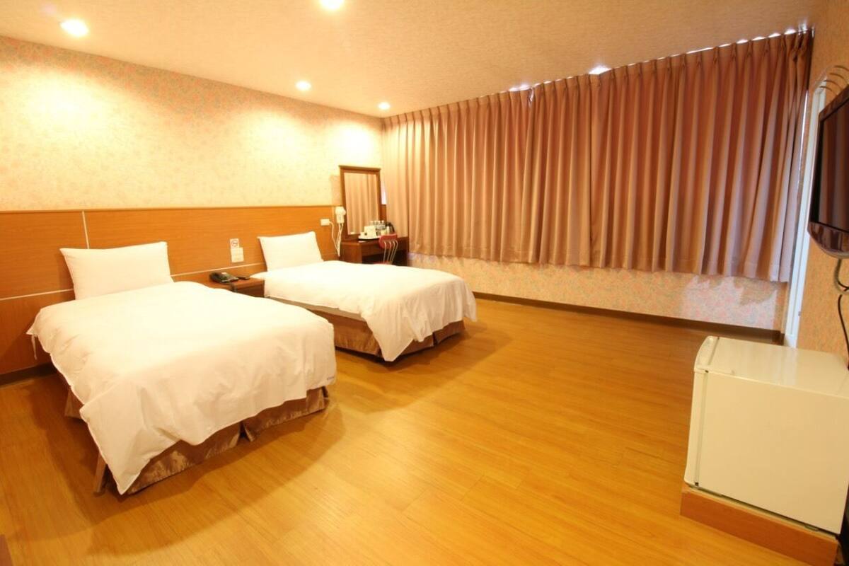 Quarto de hotel com duas camas, piso de madeira, uma janela grande com cortinas, uma televisão de tela plana e uma pequena geladeira.