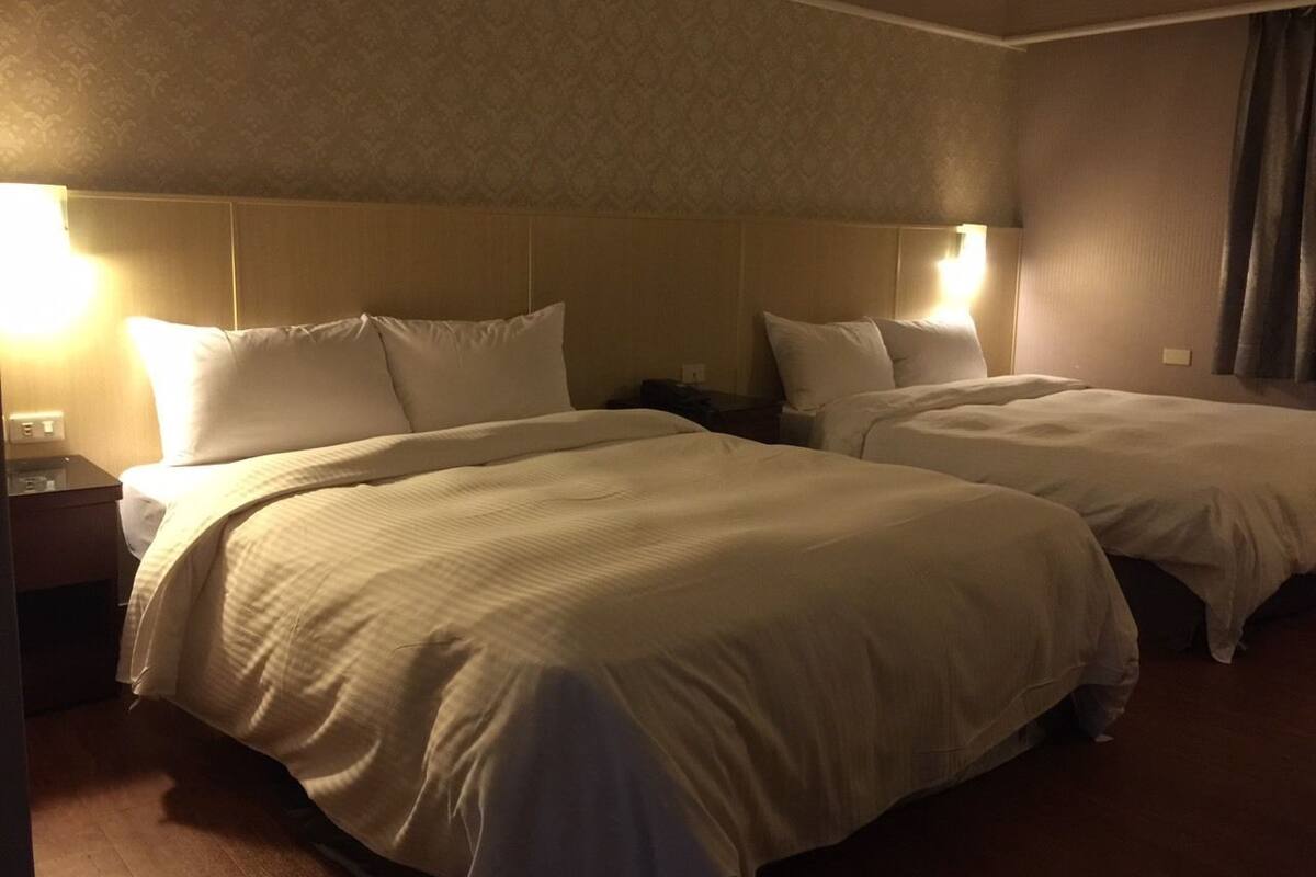 Quarto de hotel com duas camas, cada uma com roupa de cama branca e cabeceiras. Há luminárias de cabeceira e uma mesa de cabeceira com abajur.