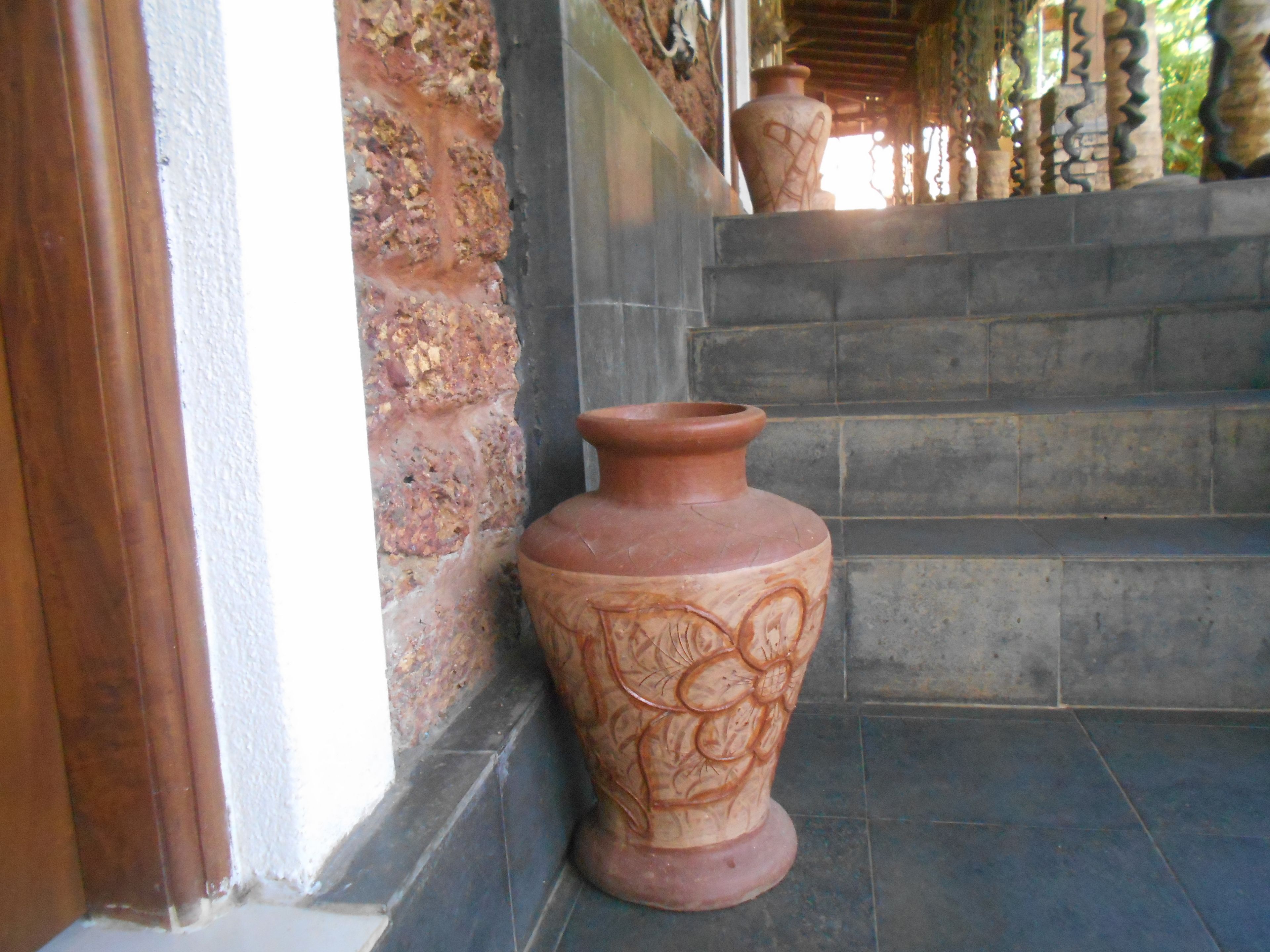 Un grand pot en terre cuite orné de motifs floraux, posé sur un sol carrelé, à côté d’un mur en pierre.