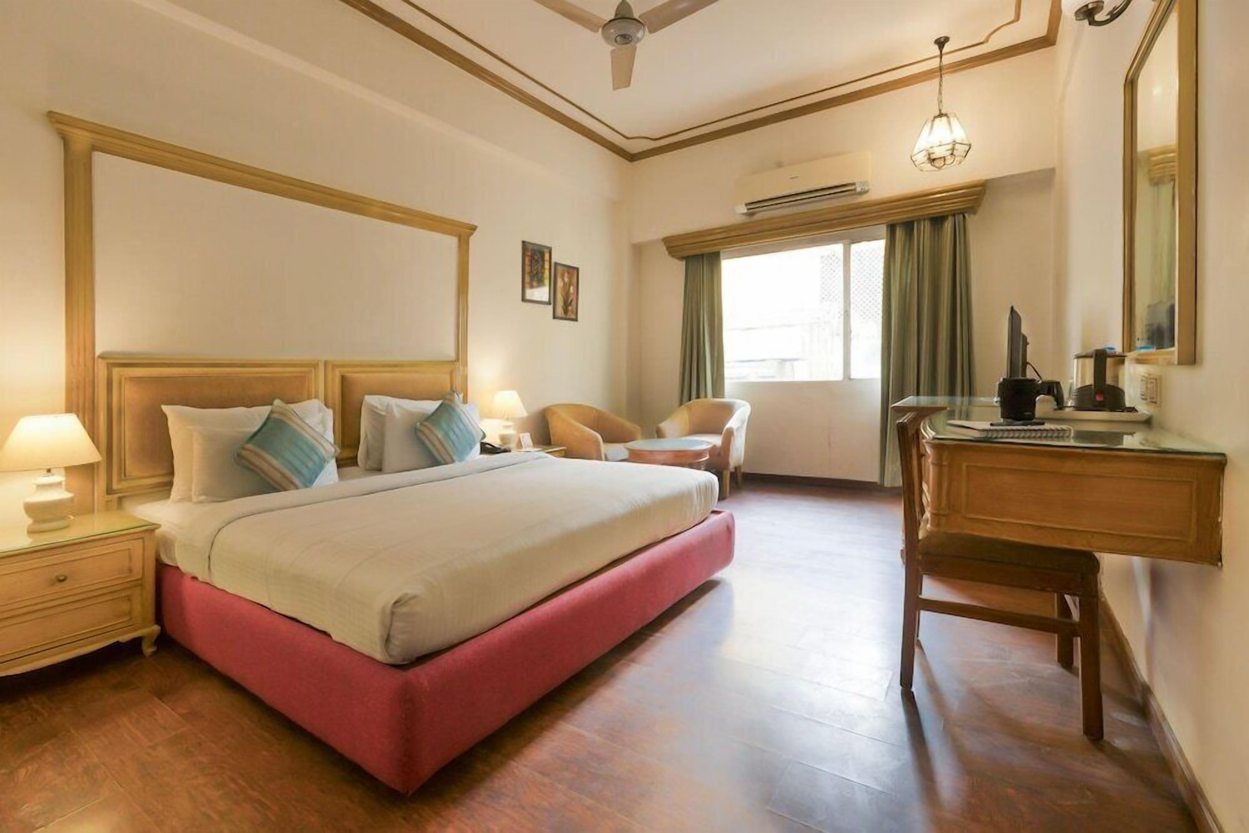 Habitación de hotel con una cama grande, un escritorio, una silla, una mesita de noche, un ventilador de techo y una ventana con cortinas.