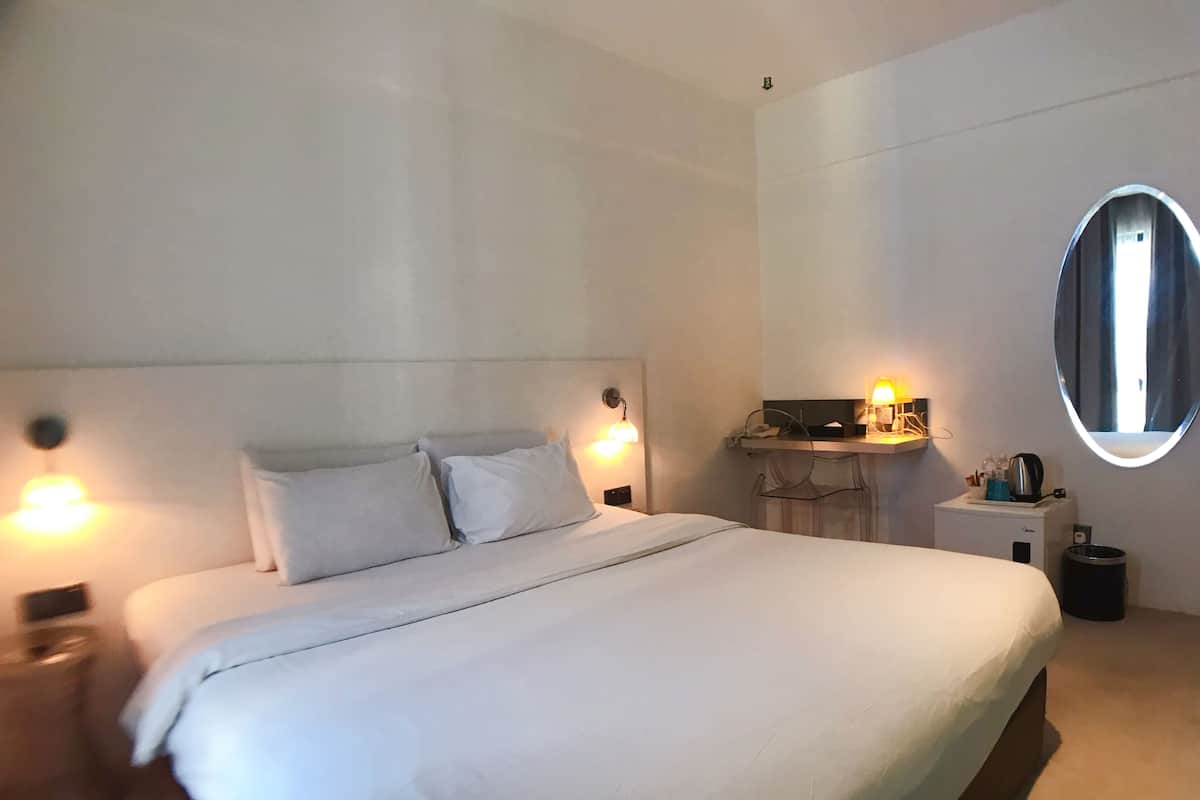 A modern hotel room with a large bed, bedside lamps, a small desk with a lamp, and a round mirror on the wall.