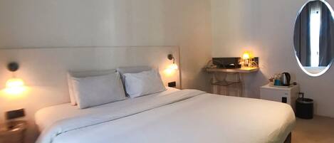 A modern hotel room with a large bed, bedside lamps, a small desk with a lamp, and a round mirror on the wall.