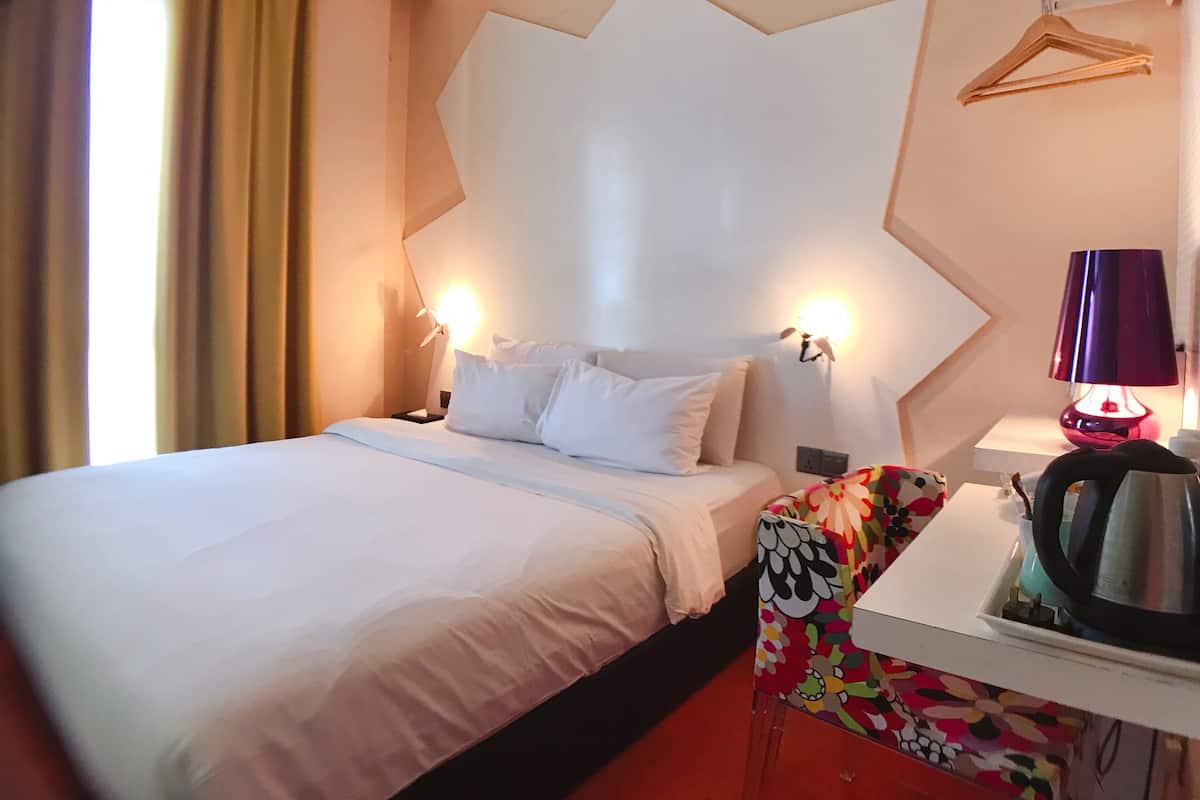 A hotel room with a bed, bedside lamps, a desk with a kettle, and a colorful chair.