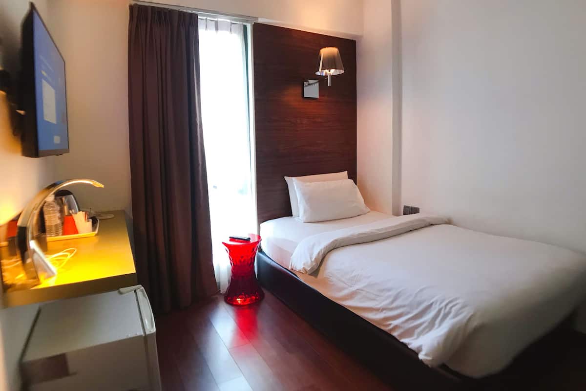 A hotel room with a bed, a desk with a lamp, a red vase, and a window with curtains.