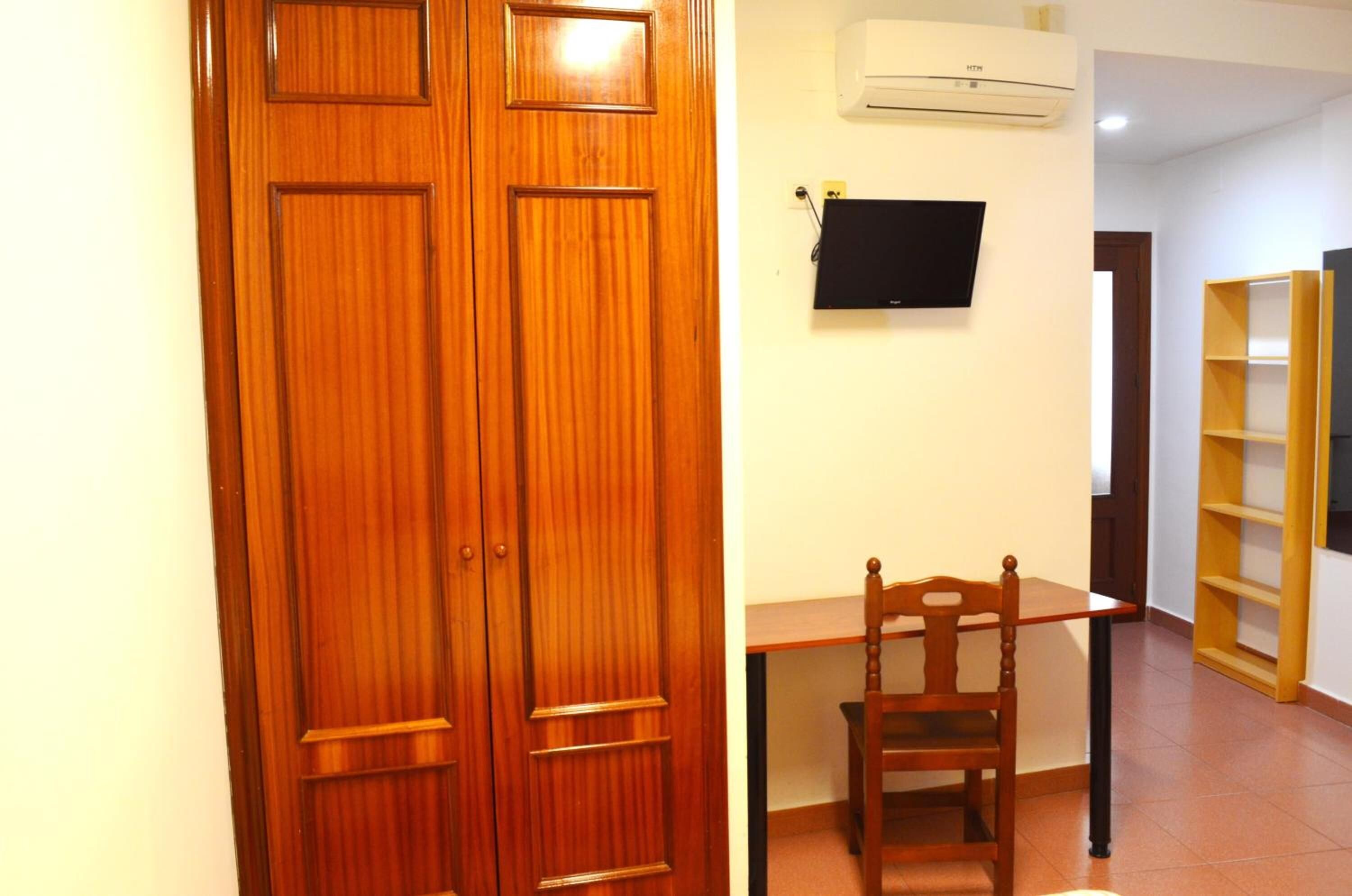 Uma porta de madeira, uma escrivaninha com uma cadeira, uma televisão e uma estante de livros num quarto.