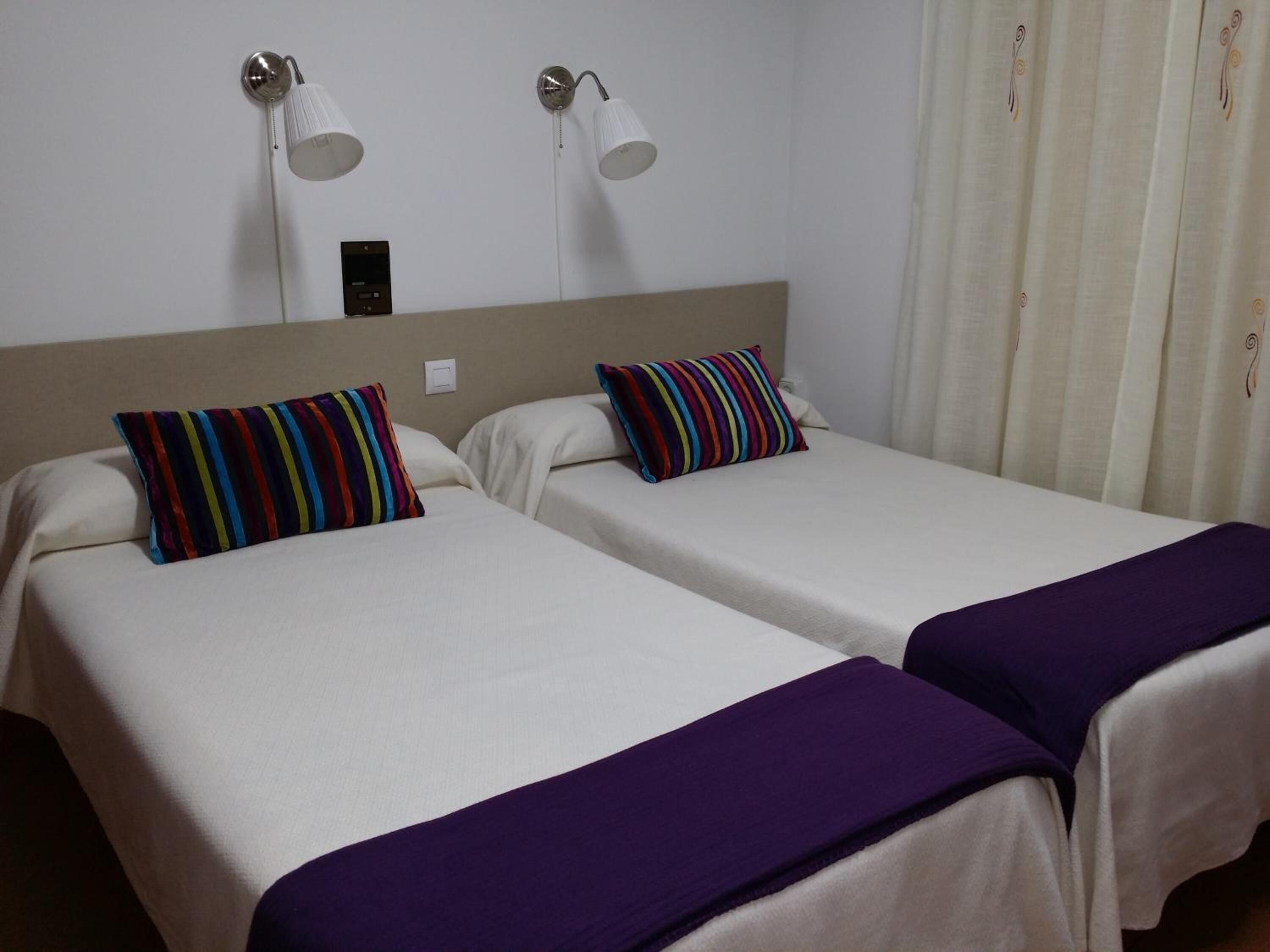 Ein Hotelzimmer mit zwei Betten, jeweils mit weißer Bettdecke und violetter Decke. Es gibt zwei Wandlampen mit weißen Lampenschirmen.