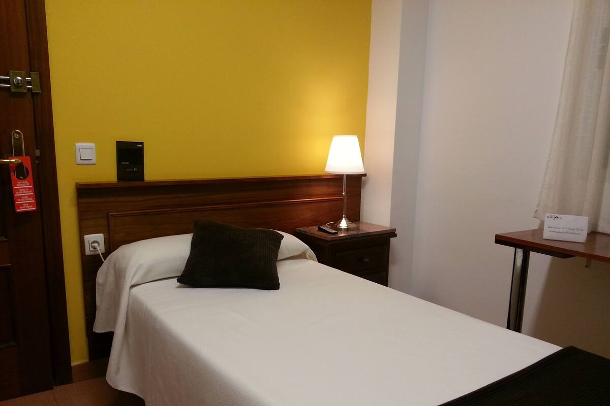 Une chambre d’hôtel comprenant un lit, une table de chevet avec une lampe, une petite table et un thermostat fixé au mur.