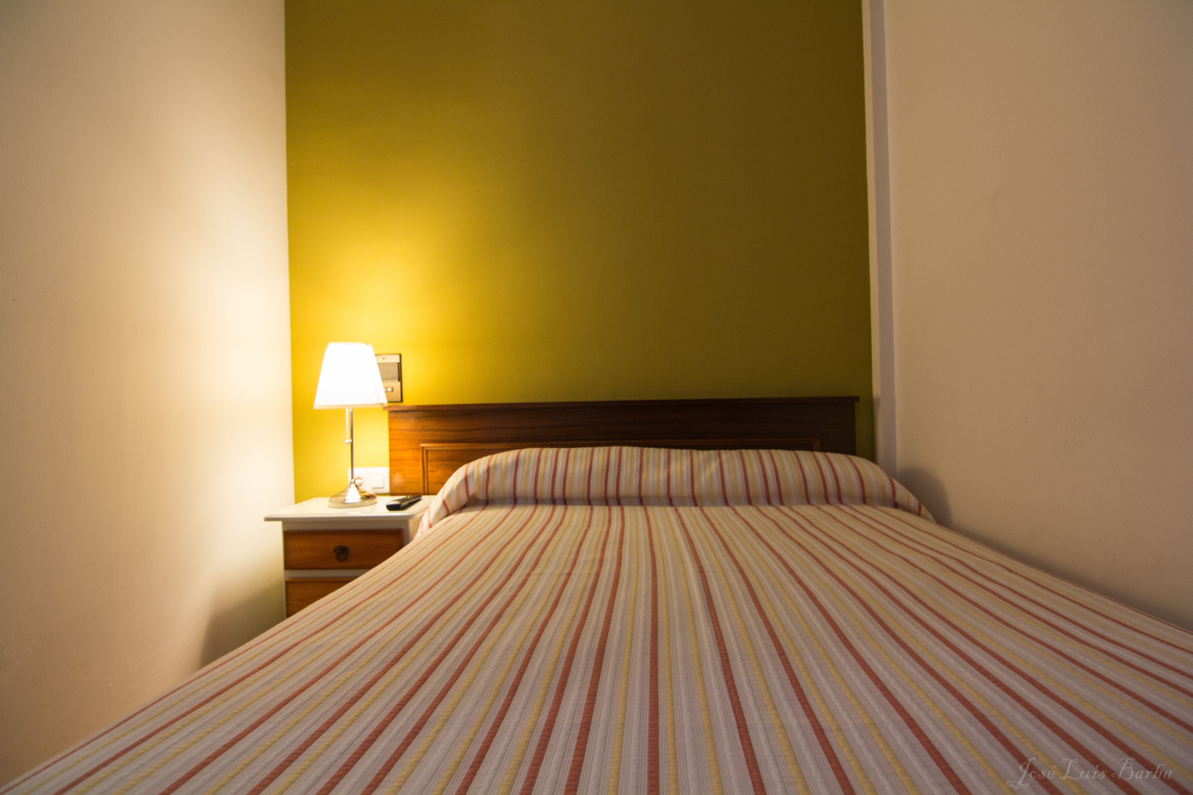 Habitación de hotel con cama, mesita de noche, lámpara y pared con un detalle amarillo.