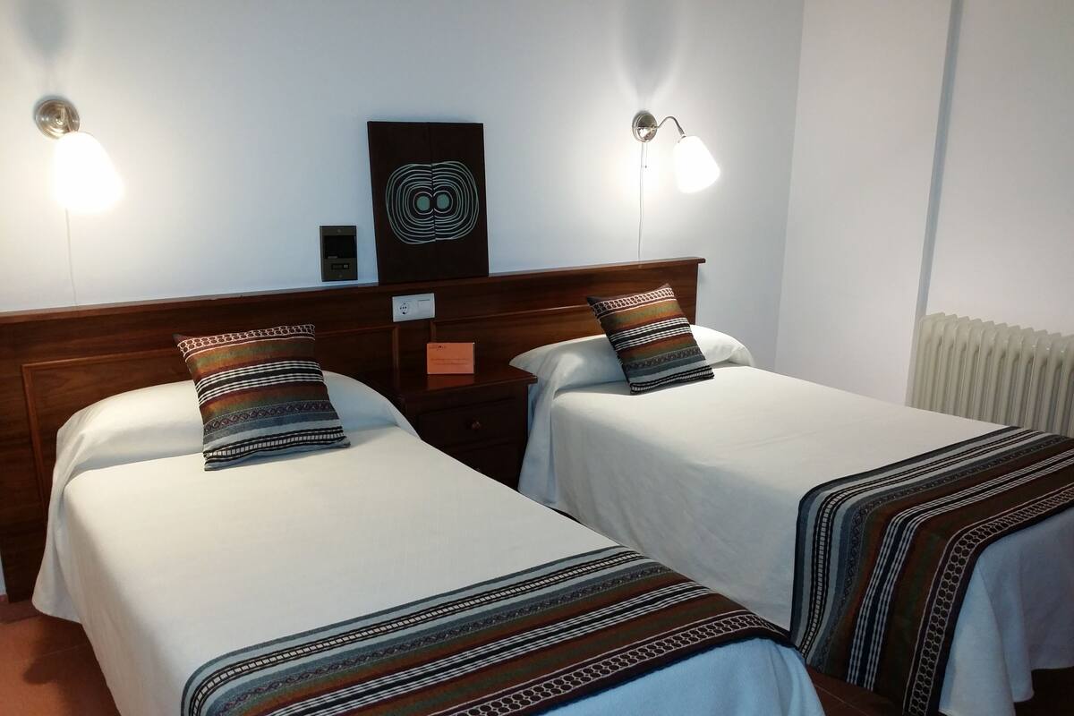 Une chambre d’hôtel avec deux lits simples, chacun agrémenté d’une couvre-lit à motifs et d’un coussin décoratif. Des lampes murales sont fixées au-dessus de chaque lit, et une petite table se trouve entre les deux.